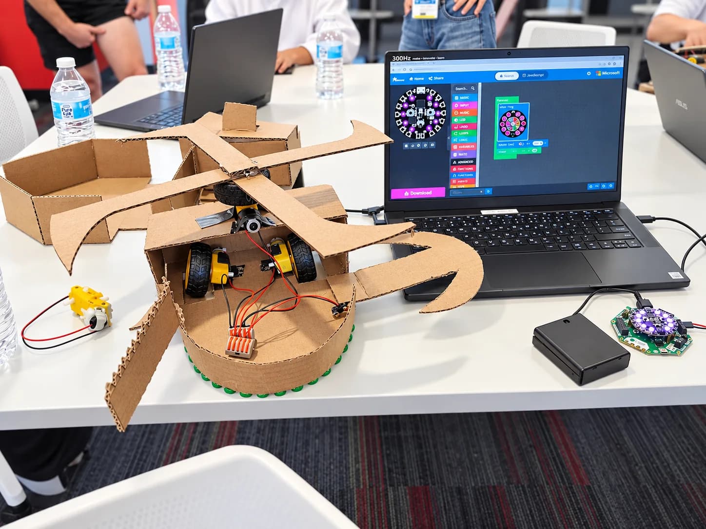 A cardboard robot beside a laptop showing visual code and a connected circuit board